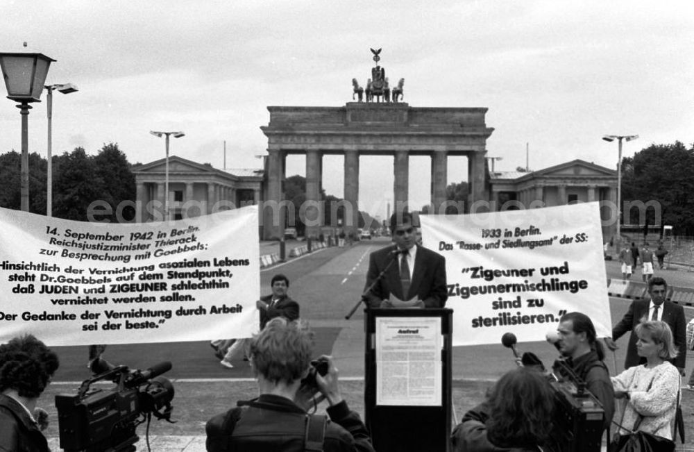 Berlin-Mitte: Sintis und Roma demonstrieren auf dem Pariser Platz vor dem Brandenburger Tor. Mann steht an Rednerput, davor Pressevertreter (Rückansicht). Demonstranten halten Plakate.
