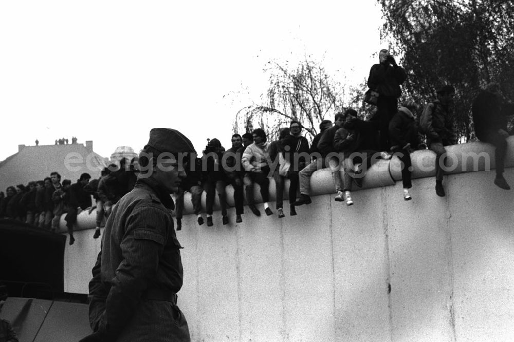 GDR picture archive: Berlin - Soldaten der DDR stehen in einer Reihe vor der Berliner-Mauer am Potzdamer Platz, auf der Mauer sitzen und stehen Menschen.