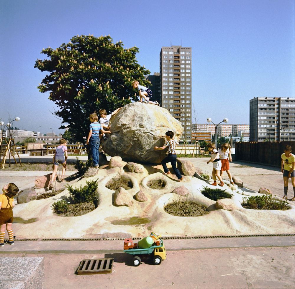 Berlin: Children's playground at Anton-Saefkow-Platz in the Lichtenberg district of EastBerlin in the territory of the former GDR, German Democratic Republic. Children are playing on a large boulder, a dump truck with digging equipment is standing in front of it