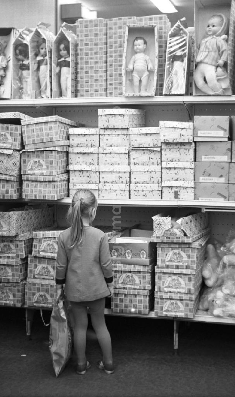 GDR picture archive: Berlin - Ob Mutti die mir kauft?, denkt dieses Mädchen vielleicht, das in der Spielwarenabteilung im Centrum Warenhaus am Alex in Berlin vor einem Regal mit Puppen steht und in einen geöffneten Karton schaut.