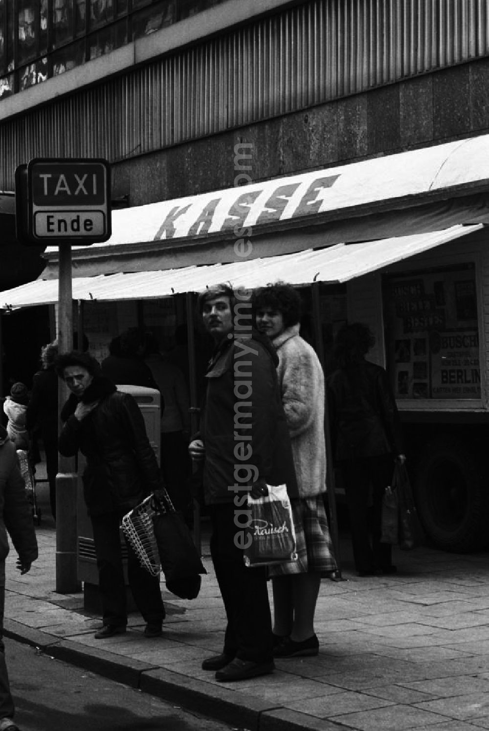 Berlin: Unweit eines Kassenwagen für den Zirkus Busch warten Menschen am S-Bahnhof Alexanderplatz in Berlin auf ein Taxi am Taxistand. Das kostet vor allem Zeit. Taxen sind auf den DDR-Straßen rar, deshalb verdienen sich auch viele private PKW-Besitzer mehr oder weniger geduldet als Schwarztaxi noch Geld hinzu.