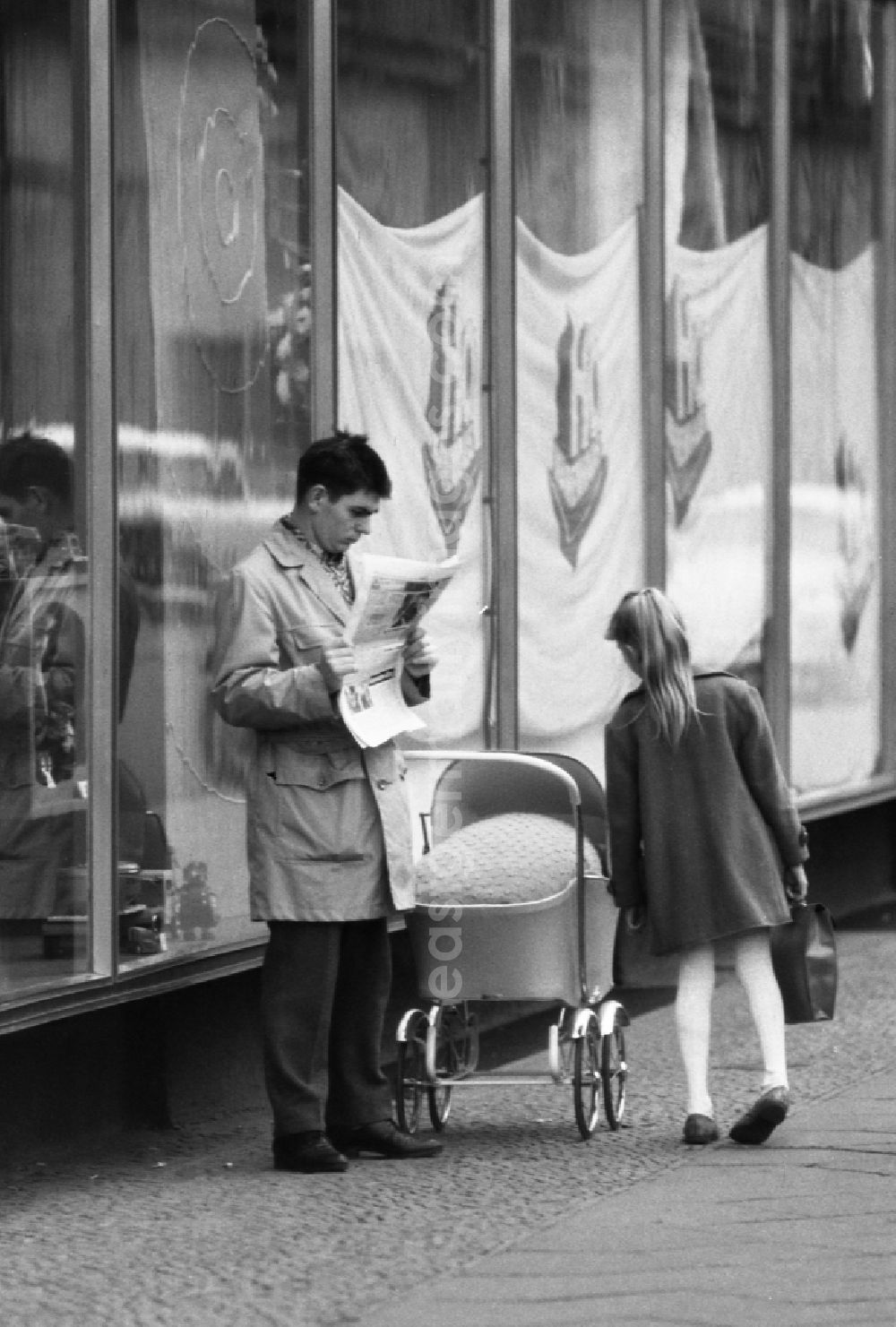 GDR photo archive: Berlin - Girl looks into the stroller of a father reading a newspaper in East Berlin on the territory of the former GDR, German Democratic Republic