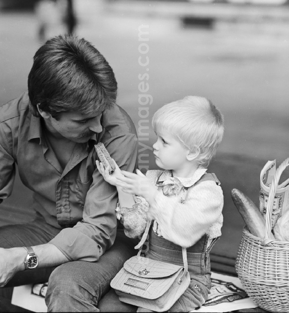 GDR photo archive: Berlin - Father sitting with his child on a bench and eat a sandwich in Berlin, the former capital of the GDR, the German Democratic Republic