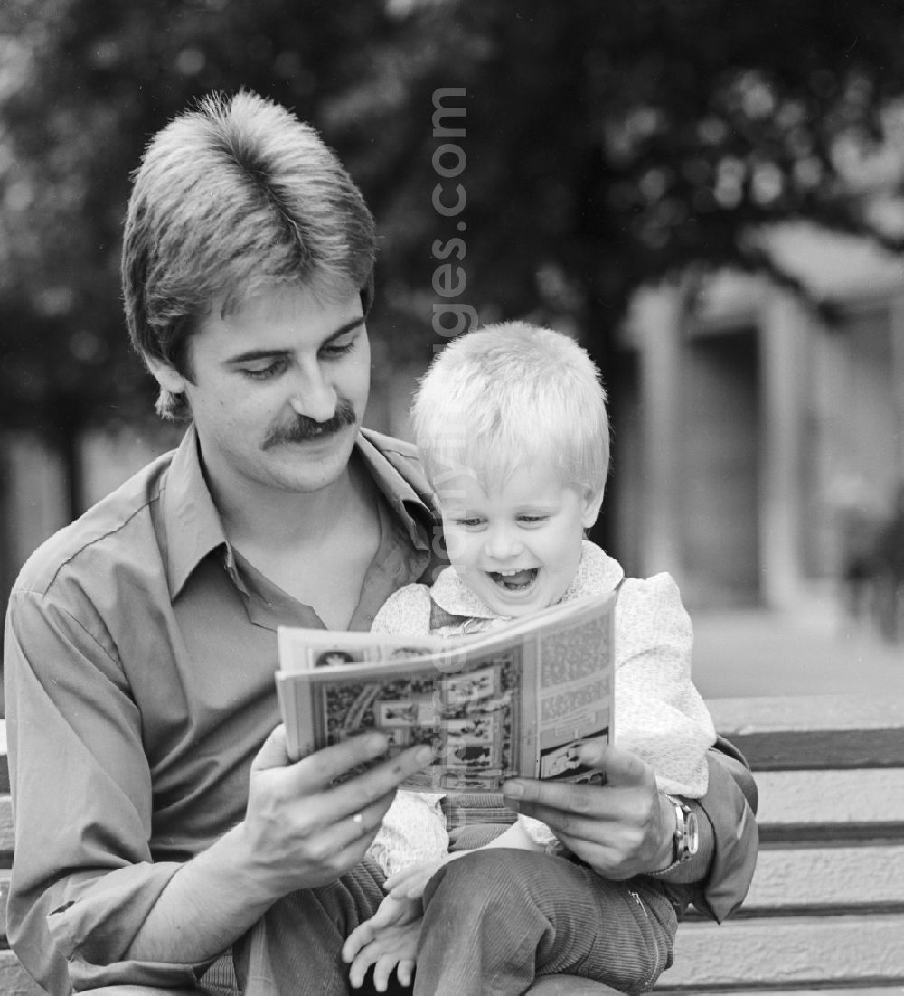 GDR image archive: Berlin - Father sitting with his child on a bench and read a magazine in Berlin, the former capital of the GDR, the German Democratic Republic