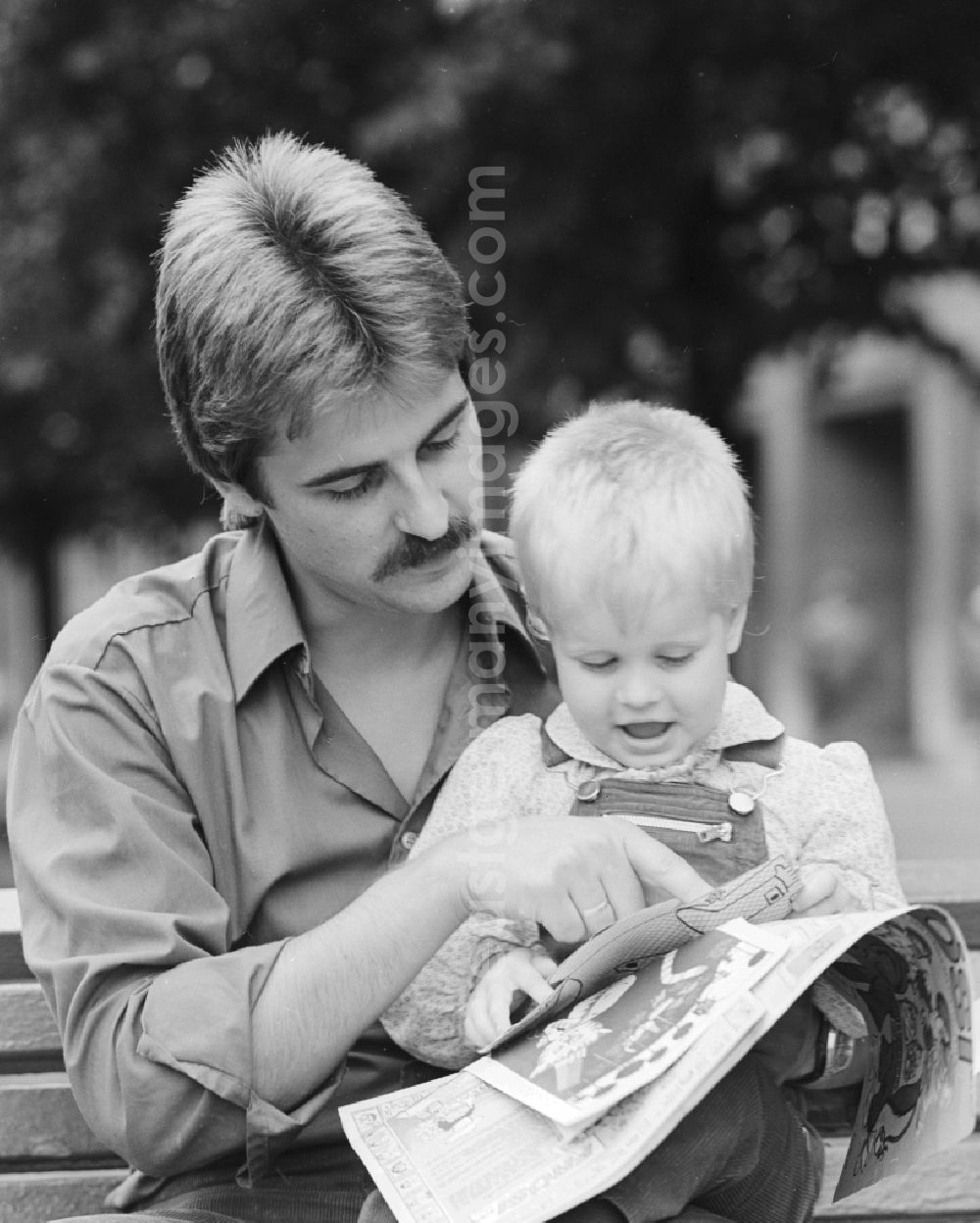 GDR picture archive: Berlin - Father sitting with his child on a bench and read a magazine in Berlin, the former capital of the GDR, the German Democratic Republic