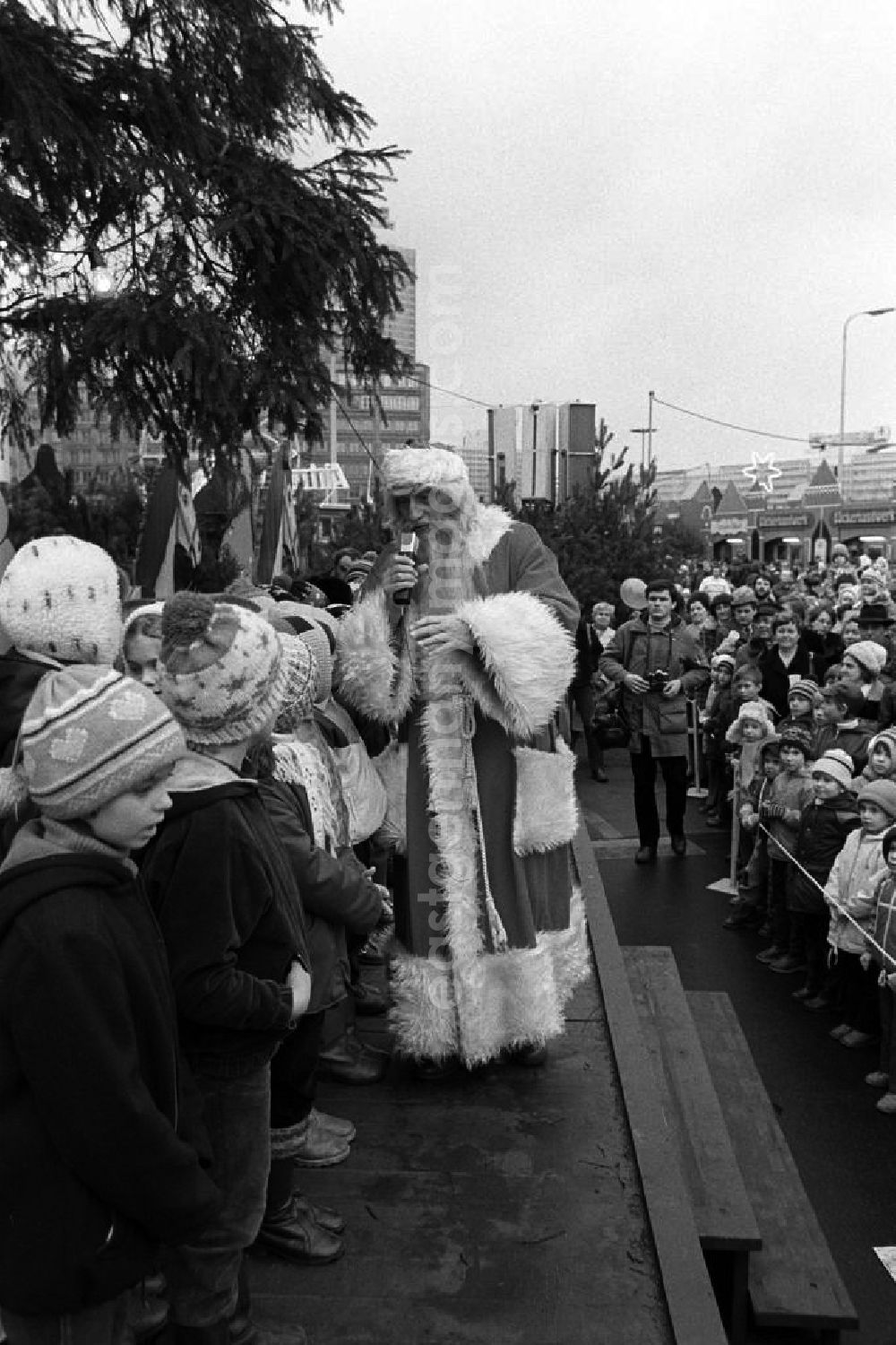 GDR picture archive: Berlin - Ein als Weihnachtsmann verkleideter Erwachsener steht auf der Bühne mit Mikrofon in der Hand vor Kindern auf dem Weihnachtsmarkt Mitte. Davor stehen Kinder hinter einem Absperrband und lauschen gebannt zu.