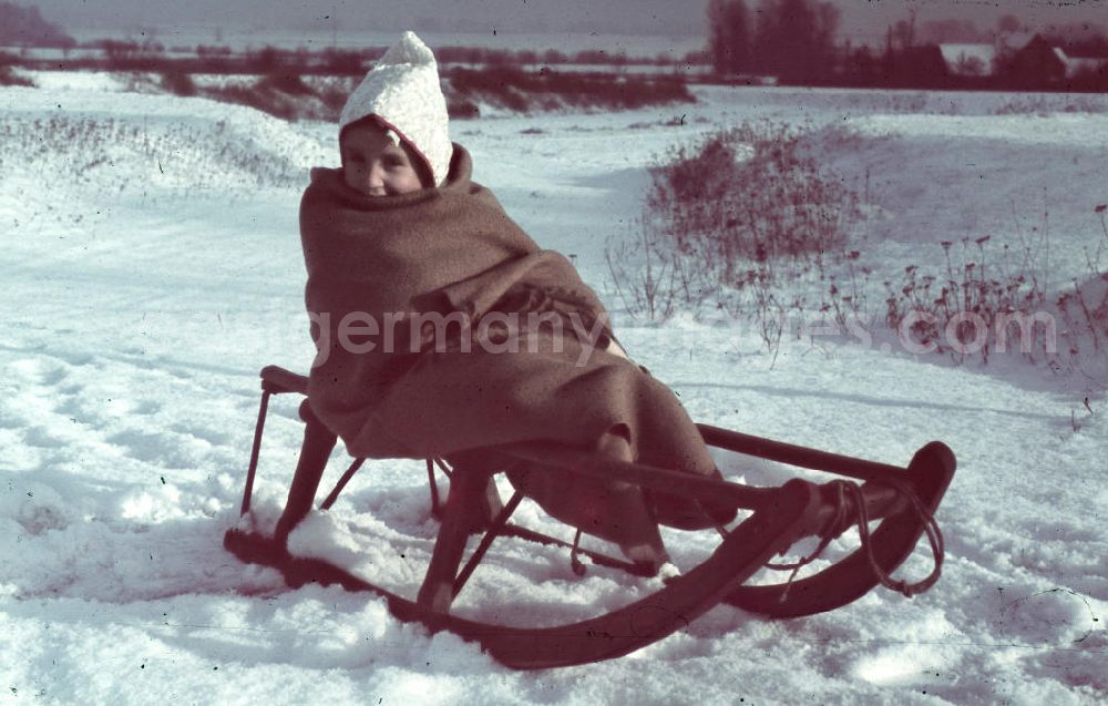 Merseburg: Kind mit Mütze sitzt in eine Decke eingewickelt auf einen Schlitten. Child with a cap sits wrapped in a blanket on a sleigh.