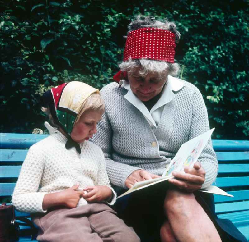 A granny sits with her grandchild on a park-bench and they look to themselves a children's book in in Ahrenshoop in the federal state Mecklenburg-West Pomerania in the area of the former GDR, German democratic republic