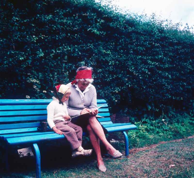 A granny sits with her grandchild on a park-bench and they look to themselves a children's book in in Ahrenshoop in the federal state Mecklenburg-West Pomerania in the area of the former GDR, German democratic republic