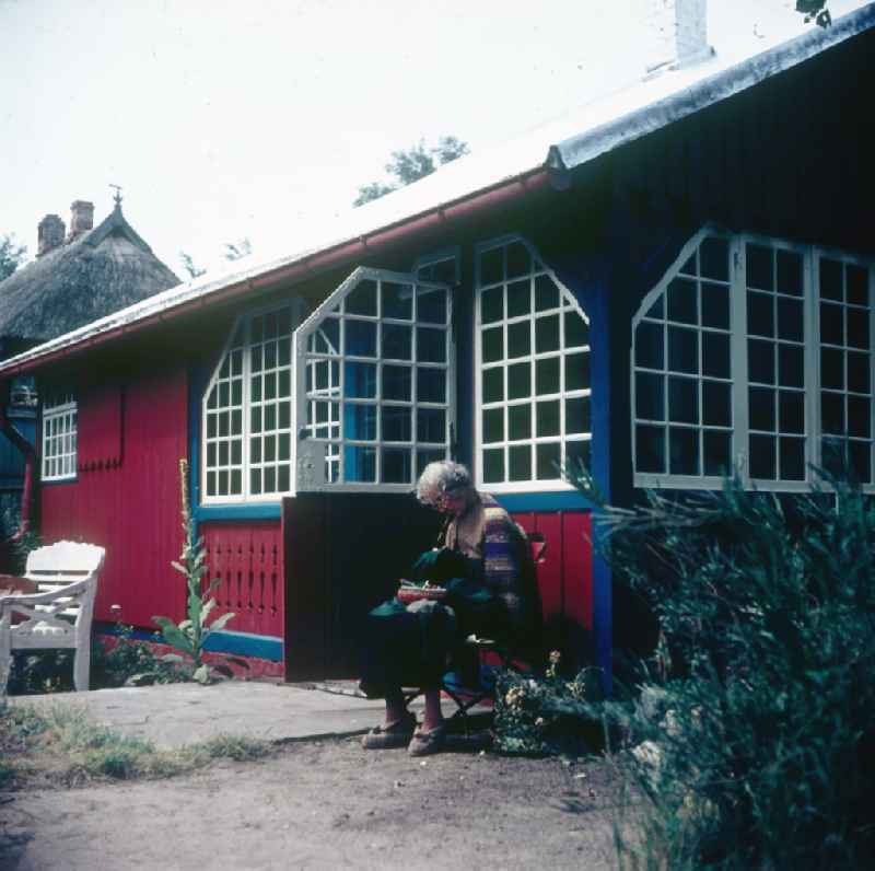 An older man sits before his red summer small house and sews in Ahrenshoop in the federal state Mecklenburg-West Pomerania in the area of the former GDR, German democratic republic