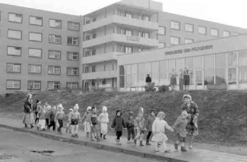 Zur Faschingszeit kostümierte Kinder laufen in Begleitung von zwei Erzieherinnen an einem Pflegeheim für Senioren vorbei. Rentnerinnen / Senioren / Omas stehen vor Pflegeheim und schauen den Kindern hinterher.