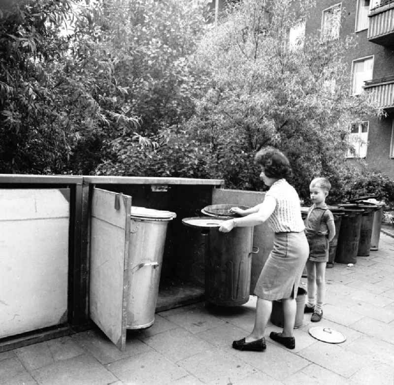 Frau steh vor Mülltonnen eines Mehrfamilienhaus, ein Junge / Kind steht daneben und schaut zu.