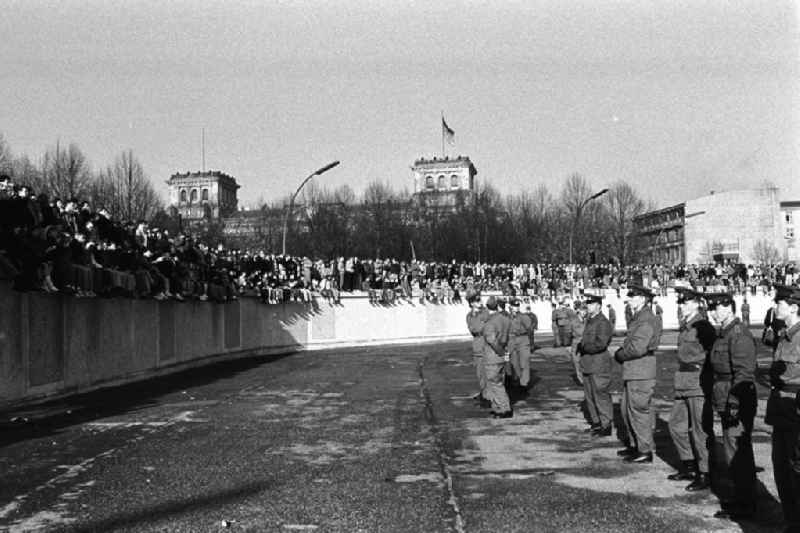 Soldaten der DDR stehen in einer Reihe vor der Berliner-Mauer am Brandenburger Tor, auf der Mauer sitzen und stehen Menschen. Im Hintergrund der Reichstag.