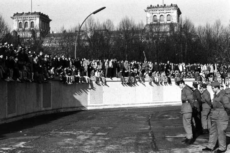 Soldaten der DDR stehen in einer Reihe vor der Berliner-Mauer am Brandenburger Tor, auf der Mauer sitzen und stehen Menschen. Im Hintergrund der Reichstag.