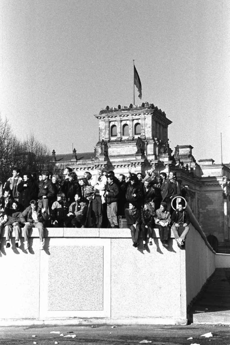 Soldaten der DDR stehen vor der Berliner-Mauer am Brandenburger Tor, auf der Mauer sitzen und stehen Menschen. Im Hintergrund der Reichstag.