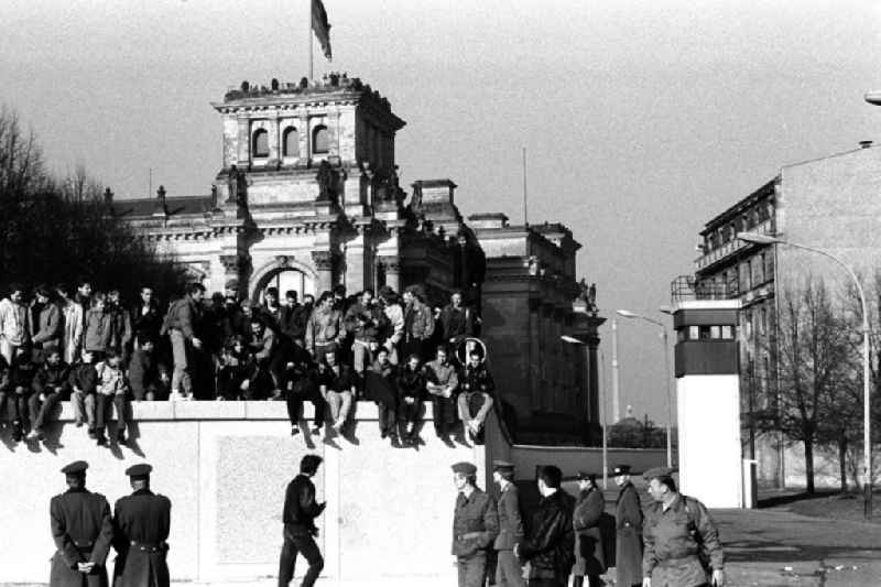 Soldaten der DDR stehen vor der Berliner-Mauer am Brandenburger Tor, auf der Mauer sitzen und stehen Menschen. Im Hintergrund der Reichstag und ein Wachturm.