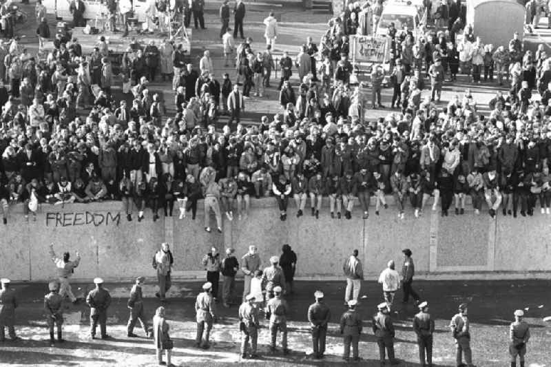 Soldaten der DDR stehen in einer Reihe vor der Berliner-Mauer am Brandenburger Tor, auf der Mauer sitzen und stehen Menschen.
