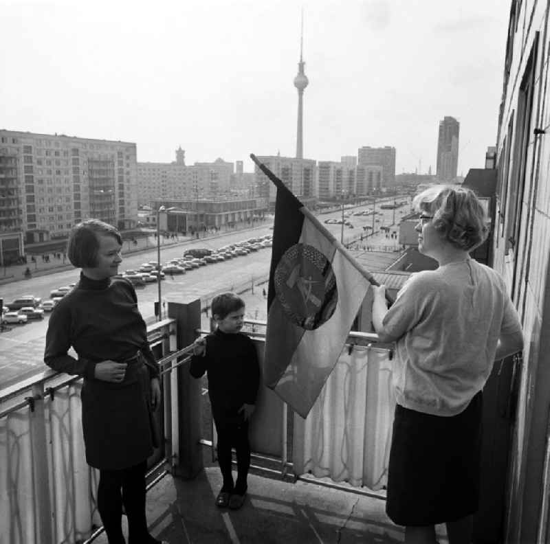 An einem Neubaublock an der Karl-Marx-Allee, der Pracht- und Paradestraße von Berlin (Ost), wird auf dem Balkon der Familie von Susanne Jessen die DDR-Fahne zum 1. Mai aufgehängt. Die Menschen, die hier zu DDR-Zeiten mit Blick auf den Alexanderplatz (Hintergrund) wohnten, gehörten zur privilegierten Schicht bzw. waren angesehene Arbeiter. An den Feiertagen, wie 1. Mai oder 7.Oktober, wurde unten auf der achtspurigen Straße die Tribüne für die Partei- und Staatsführung aufgebaut, auf der die Politiker dann huldvoll die vorbeiziehenden 'Werktätigen' begrüßten.