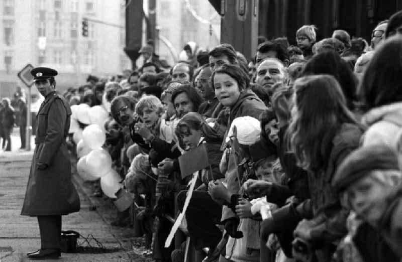 Die Bevölkerung / Menschen stehen hinter Absperrung am Straßenrand zusammen und warten auf die bevorstehende Militärparade auf der Karl-Marx-Alle anlässlich des 25-jährigen Bestehen der DDR. Kinder halten Fahnen / Winkelemente in ihren Händen. Ein Volkspolizist mit Uniform steht vor der Absperrung.