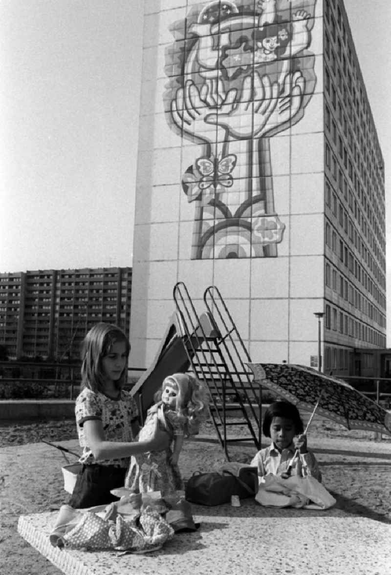 Mädchen spielen mit Puppen auf einem Spielplatz an einem Neubaugebiet in Berlin. Ein Mädchen hält eine Schirm in der Hand. An der Fassade des elfgeschossigen Wohnhauses im Hintergrund ist ein großflächiges Bild angebracht, das schützende Hände und das Glück der Kinder auf der ganzen Welt symbolisiert.