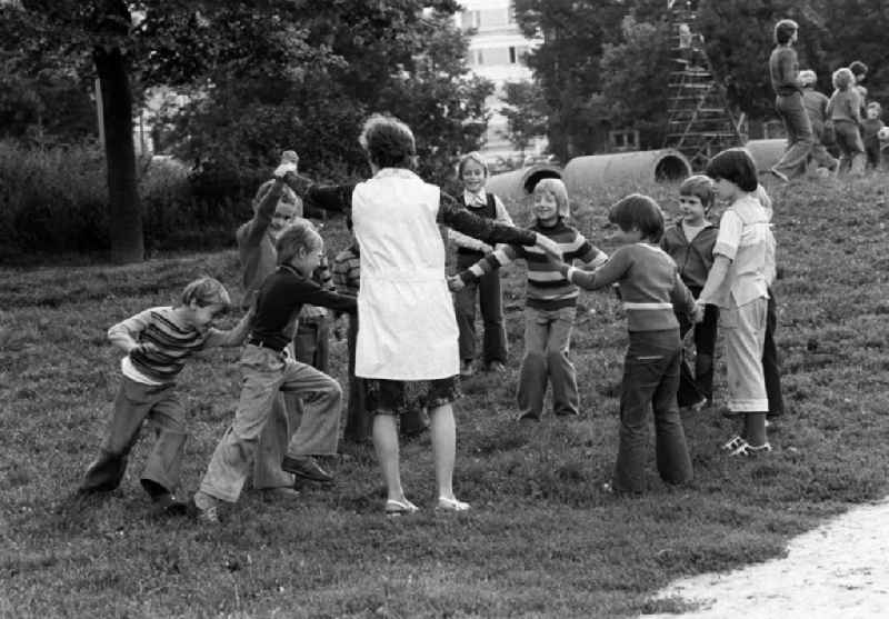 Kinder spielen auf einer Wiese neben einem Spielplatz im Neubaugebiet mit einer Erzieherin ein Kreisspiel.
