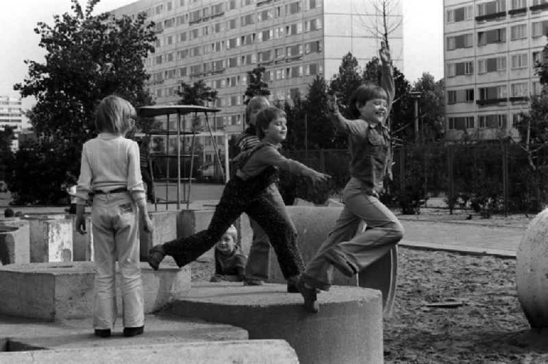 Kinder toben und spielen auf einem Spielplatz an einem Neubaugebiet in Berlin. Im Hintergrund stehen zwei zehngeschossige Plattenbauten.