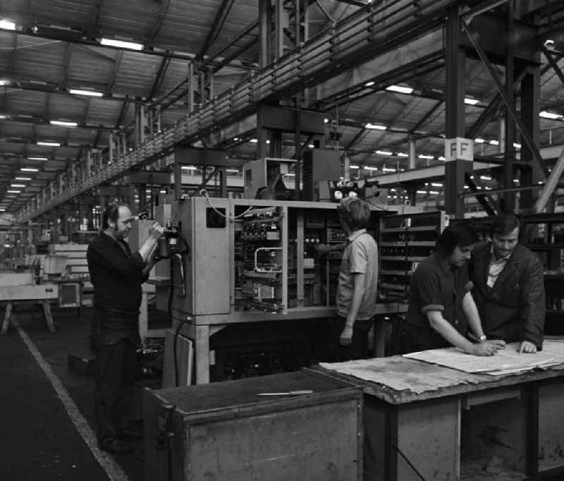 Arbeiter während der Produktion von Werkzeugmaschinen in der VEB Berliner Werkzeugmaschinenfabrik (BWF) Marzahn. Zwei Arbeiter arbeiten an einer Maschine, zwei weitere stehen zusammen vor einer technischen Zeichnung.