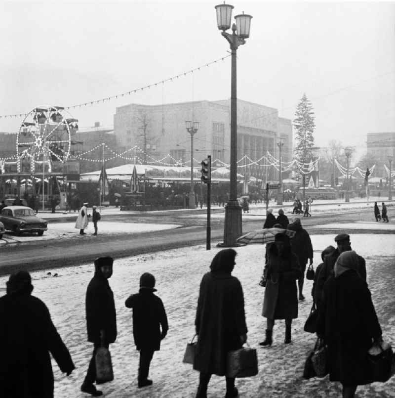 Blick über die Karl-Marx-Allee auf den Weihnachtsmarkt am Strausberger Platz. Ein Riesenrad, ein mit Lampen geschmückter Weihnachtsbaum und  aufgestellte Buden vor der Sporthalle bilden den Weihnachtsmarkt. Passanten eilen bei Schneetreiben über die Gehwege.
