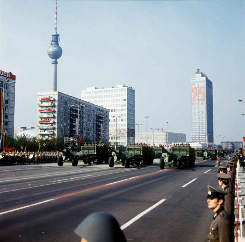 Parade der NVA auf der Karl-Marx-Allee zum 25. Jahrestag der DDR. Soldaten in Mannschaftsfahrzeugen ziehen Artilleriegeschütze. Soldaten säumen Straße im Vordergrund. Zuschauer mit Winkelementen im Hintergrund. Im Hintergrund das Haus des Lehrers, Fernsehturm, Interhotel und Centrum Warenhaus.