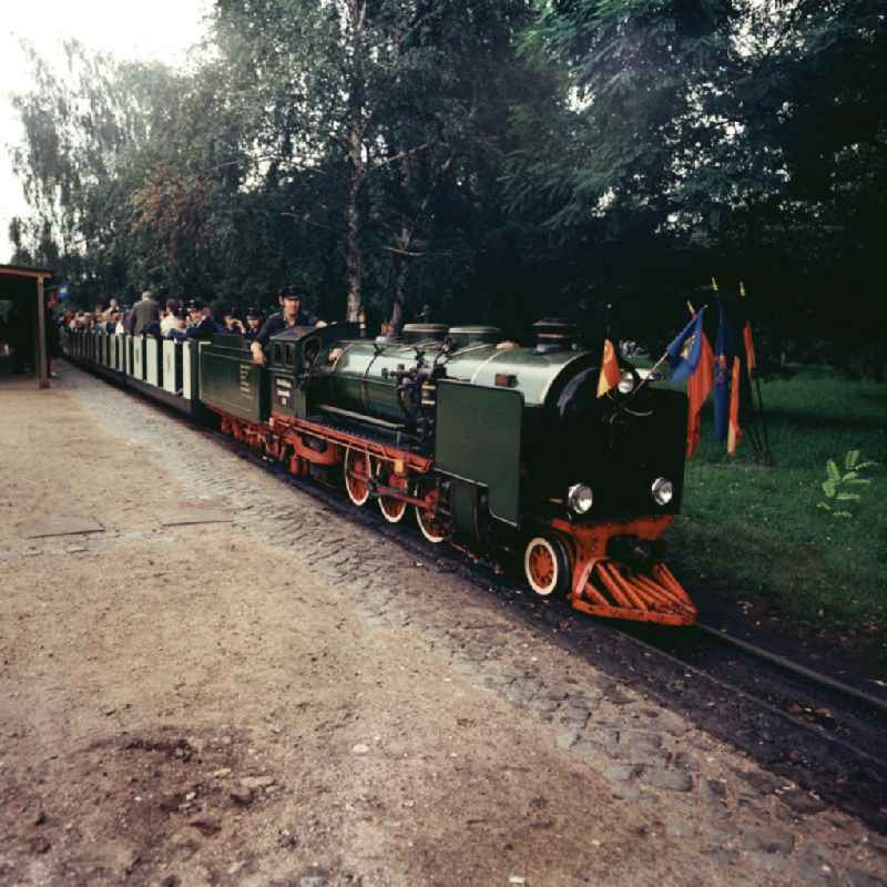 Die Pioniereisenbahn steht an einem Bahnhof / Haltepunkt in der Wuhlheide / im Pionierpark. Die Lokomotive ist mit Flaggen der DDR und der Pionierflagge geschmückt.