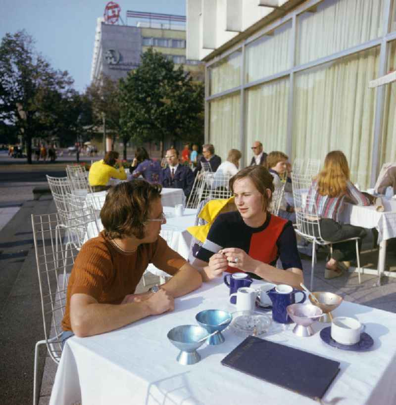 Junge Leute sitzen im Café am Lindencorso an der Friedrichstraße / Ecke Unter den Linden in Berlin-Mitte und unterhalten sich, auf dem Tisch stehen die typischen DDR-Eisbecher und -Geschirr. Im Hintergrund das Interhotel 'Unter den Linden'. Das Lindencorso war ein 1966 eröffneter fünfgeschossiger Gaststätten- und Bürokomplex mit vorgelagerten Terrassen im Fußgängerbereich. Beide Gebäude entsprachen den zeitgenössischen Ideen der sozialistischen Architektur. Sie wurden nach der Wende 199
