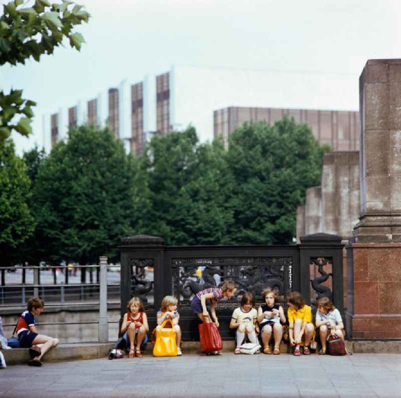 Eine Kindergruppe sitzt am Geländer der Marx-Engels-Brücke (heute Schloßbrücke) gegenüber vom Zeughaus in Berlin. Den gusseisernen Brückenschmuck zieren mythologisch-maritime Motive nach Schinkels Entwürfen, auf den roten Granitsockeln mit den hohen Marmor-Postamenten fehlen noch die Brückenfiguren. Diese wurden erst 1983 nach ihrer Restaurierung wieder aufgestellt. Im Hintergrund der Palast der Republik.