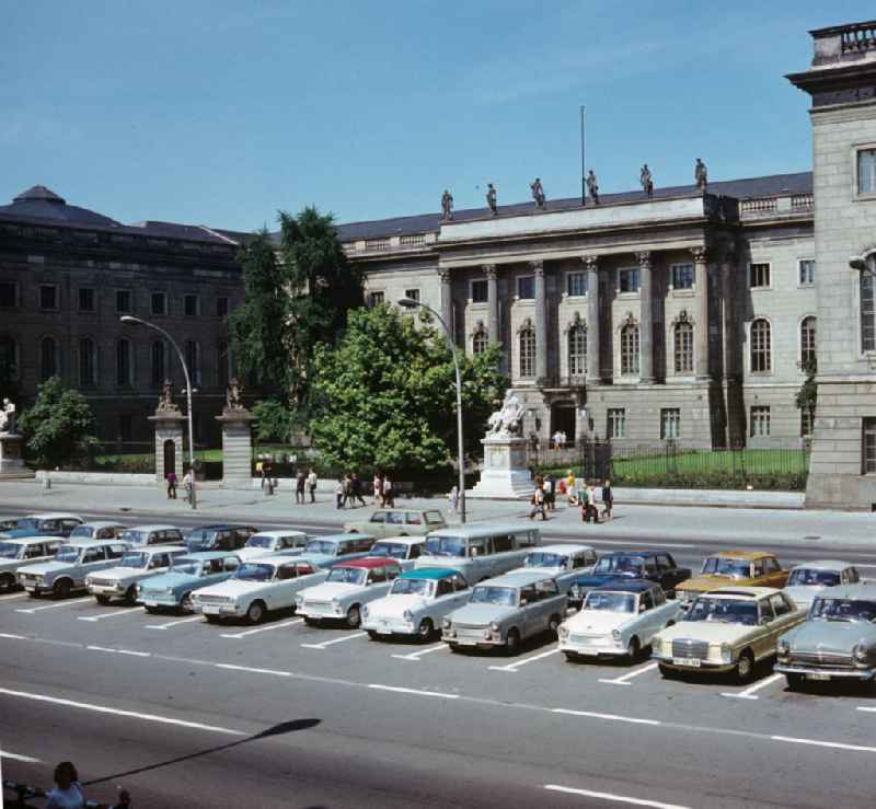 Blick auf die größte Universität der DDR, die Humboldt-Universität zu Berlin. Auf dem Mittelstreifen Unter den Linden parken überwiegend Trabbis und Wartburgs, dazwischen steht ein westdeutscher Mercedes.