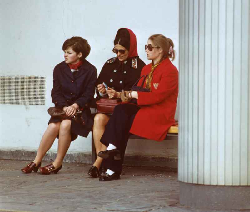 Drei schick gekleidete Frauen sitzen auf einer Bank im Säulengang am Haus des Lehrers am Alexanderplatz in Berlin-Mitte.