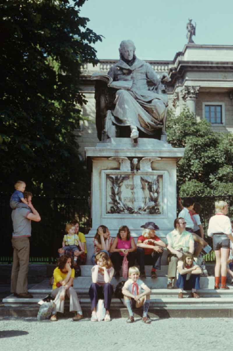 Junge Pioniere und Touristen sitzen auf den Stufen des Denkmals für Wilhelm von Humboldt, das vor dem Hauptgebäude der Humboldt-Universität Unter den Linden in Berlin steht.