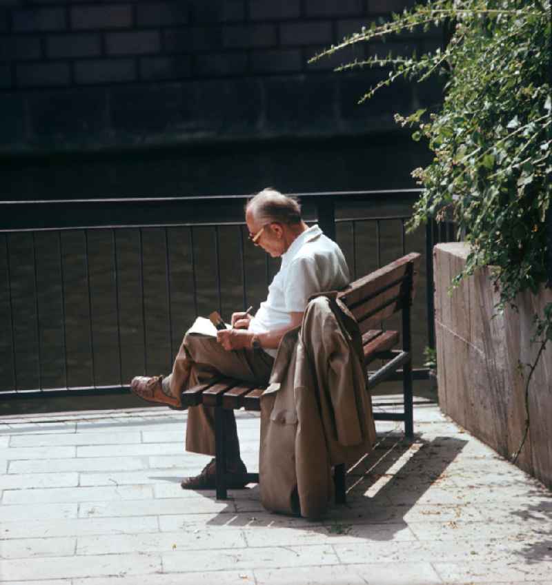 Ein Mann sitzt auf einer Parkbank am Spreeufer und löst in einer Zeitung Kreuzworträtsel.