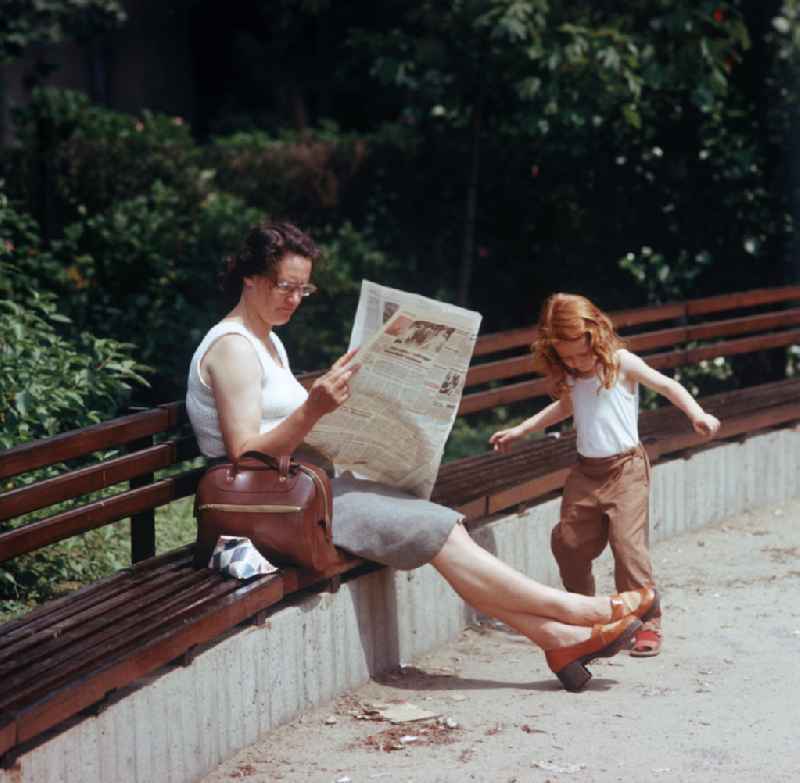 Eine Frau liest auf einer Parkbank ihre Zeitung, während ihre Tochter daneben spielt. Auf der Bank liegt ein Tetra Pak mit Trink-Vollmilch.