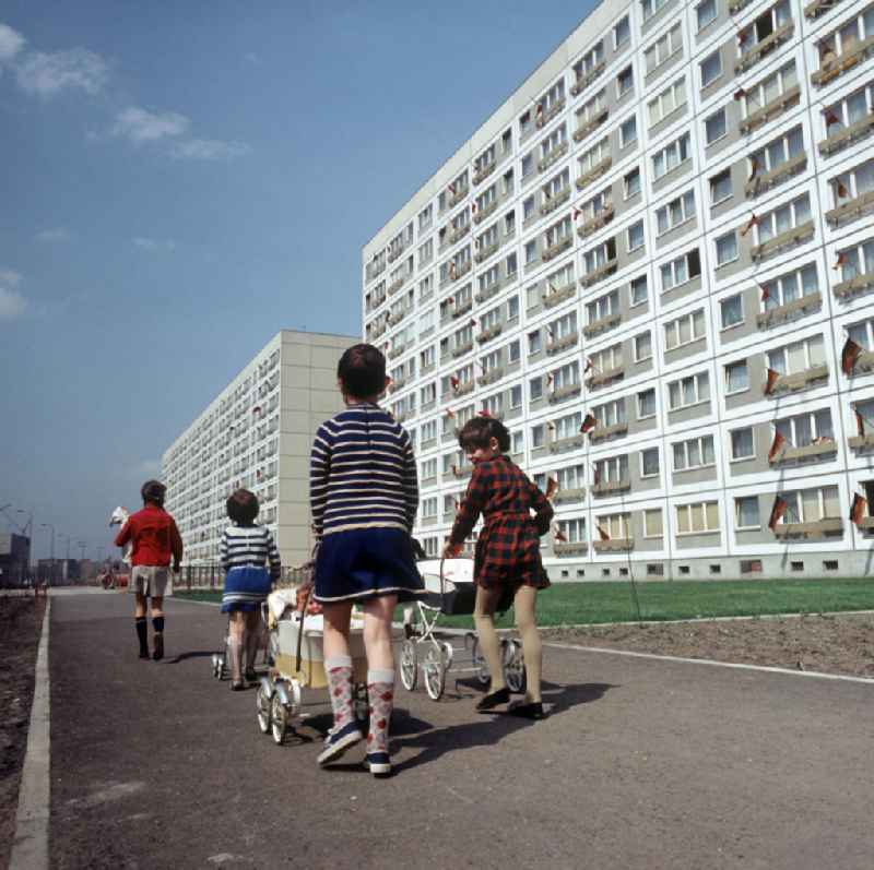 Kinder gehen mit ihren Puppenwagen spazieren, aufgenommen im Neubauviertel an der Spandauer Straße / Ecke Karl-Liebknecht-Straße. Die Häuser sind zum Maifeiertag mit Fahnen festlich geschmückt. Der bestehende Wohnungsmangel entwickelte sich in der DDR seit den 6