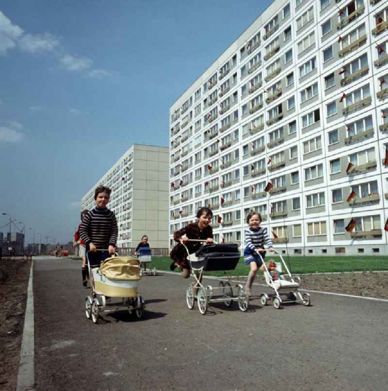Kinder machen mit ihren Puppenwagen ein Wettrennen, aufgenommen im Neubauviertel an der Spandauer Straße / Ecke Karl-Liebknecht-Straße. Die Häuser sind zum Maifeiertag mit Fahnen festlich geschmückt. Der bestehende Wohnungsmangel entwickelte sich in der DDR seit den 6