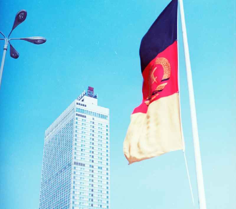 Flag of the German Democratic Republic in Berlin. It shows the colors of the Weimar Republic. On the colors black, red and gold is in the middle the coat of arms of the GDR with hammer, compasses and wreath of grain ears. In the background is the Hotel Stadt Berlin and the Berlin coat of arms, the Berlin Bear