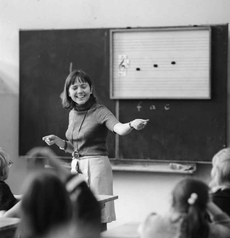 A teacher stands in front of the class and teaches music in Berlin. On the blackboard are staves with clef and notes