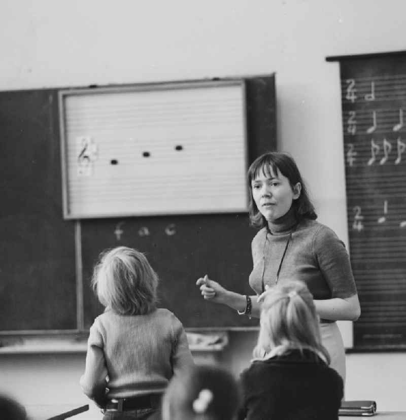 A teacher stands in front of the class and teaches music in Berlin. On the blackboard are staves with clef and notes