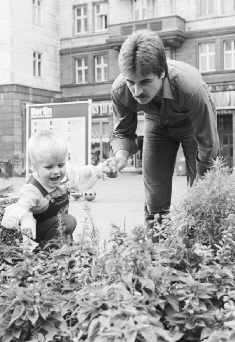 A father is holding his child in front of a flower bed in the Karl-Marx-Allee in Berlin, the former capital of the GDR, the German Democratic Republic