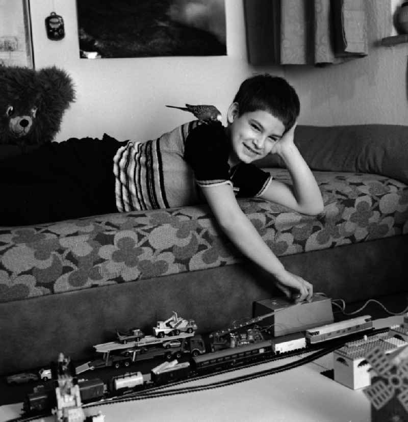 A boy playing with his model railway in his nursery in Berlin, the former capital of the GDR, the German Democratic Republic. His pet, a budgie, sitting on his back and looks on