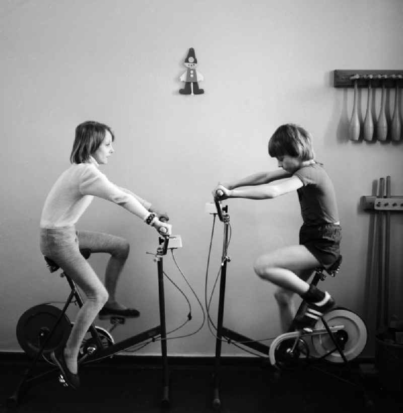 2 children exercising on exercise bikes fitness and stamina in the Children's Clinic in Klinikum Berlin-Buch in Berlin, the former capital of the GDR, the German Democratic Republic. A mother sitting with her baby sits treatment rooms