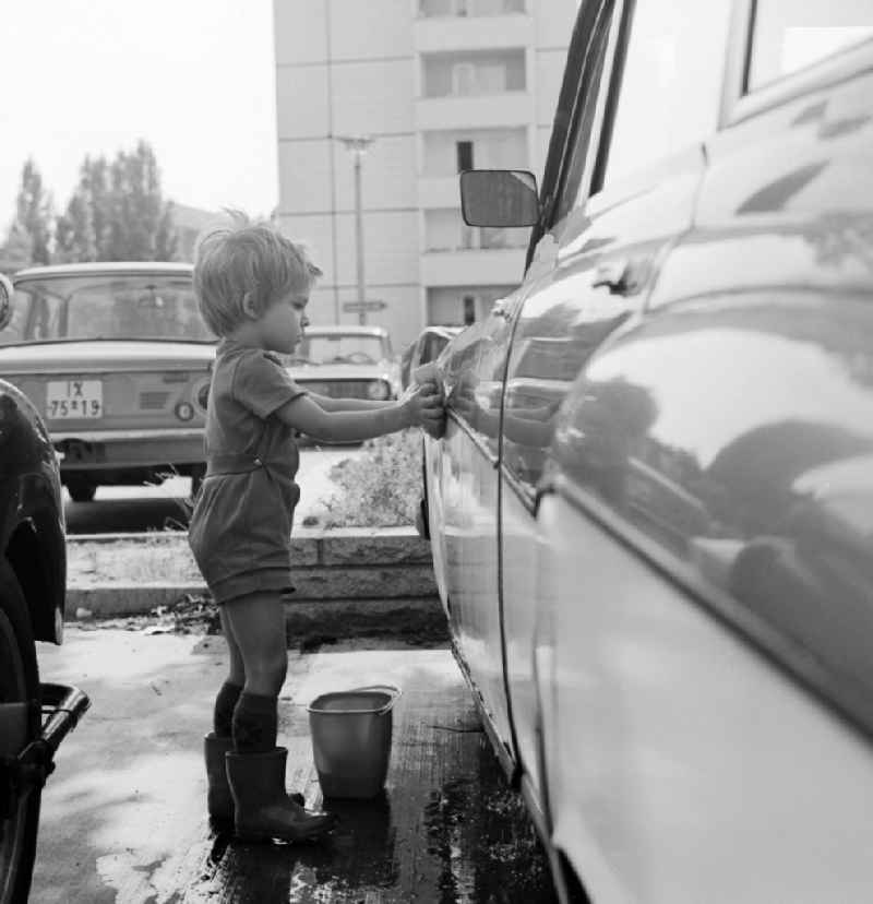 A toddler with rubber boots and bucket at the car wash in a parking lot in Berlin, the former capital of the GDR, the German Democratic Republic
