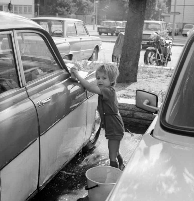 A toddler with rubber boots and bucket at the car wash in a parking lot in Berlin, the former capital of the GDR, the German Democratic Republic