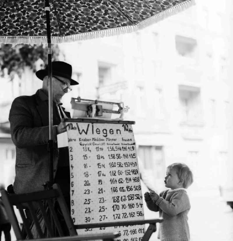 With hat and suit dressed elderly gentleman in weighing of road passersby on the Schoenhauser Allee in Berlin, the former capital of the GDR, German Democratic Republic. A small child looks reverently on the weighing table in front of the scale. For a mite of 1