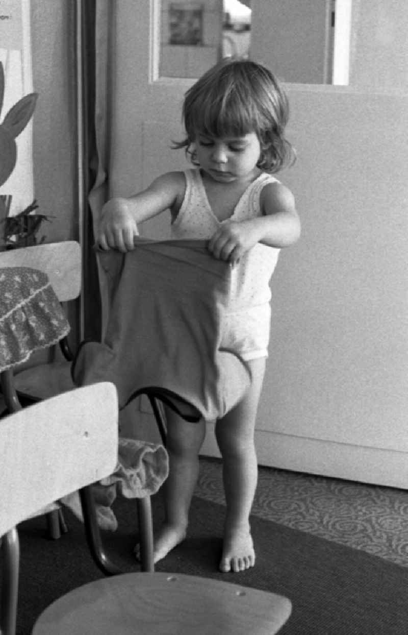 Preparing for a nap in the kindergarten in Berlin Eastberlin on the territory of the former GDR, German Democratic Republic. Toddler tries to undress on his own and pulls on his sock or stocking. Toddler undresses and puts a T-shirt over a chair