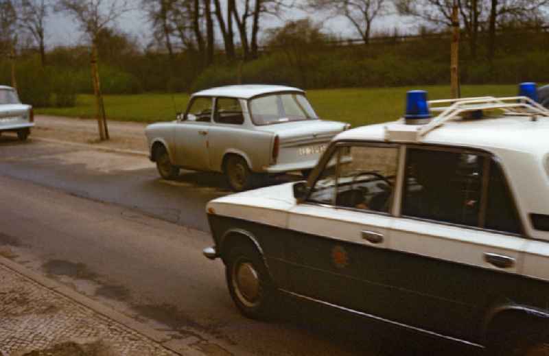 People's Police car drives on a street in East Berlin in the territory of the former GDR, German Democratic Republic. Trabant cars are parked on the side of the road